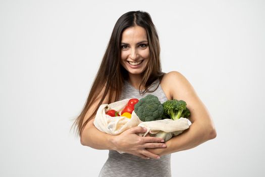 A white mesh mesh bag with groceries in a hands of a girl in grey dress against the white background of a studio