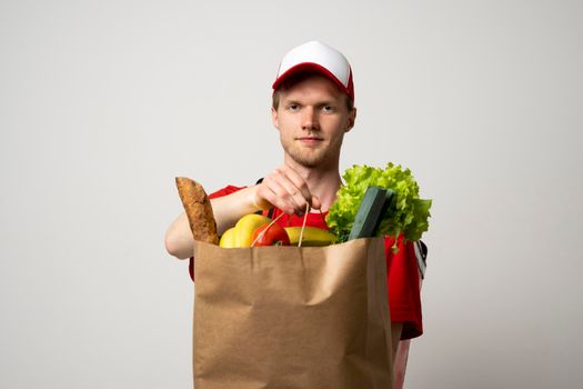 Young man in a red t-shirt and a cap holding paper bag with products on white background. Food delivery service