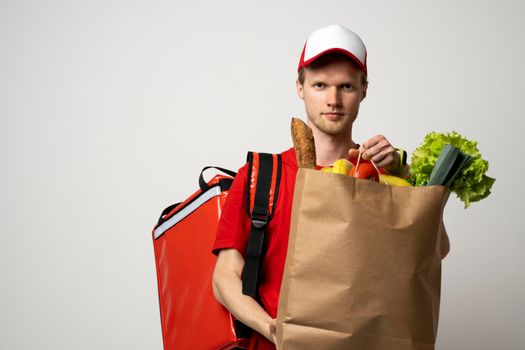 Delivery Concept. Portrait of pleased delivery man in red uniform smiling while carrying paper bag with food products isolated over white background.