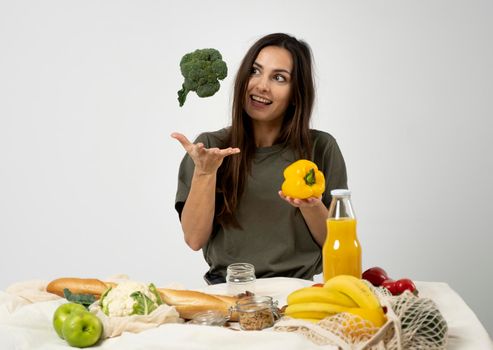 Happy woman in green t-shirt unpacking shopping mesh eco bag with healthy vegetables, fruits, bread, snacks on the kitchen at home and throwing broccoli in air. Healthy eating vegetarian concept