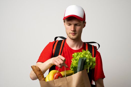 Grocery delivery courier man in a red cap and t-shirt with a thermal bag on a shoulders holds paper bag with food on white studio background. Food delivery service, online shopping