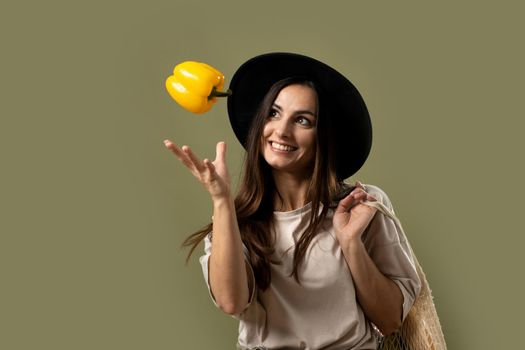 Smiling brunette young woman in a beige t-shirt and a hat holding a reusable mesh bag with fresh vegetables and fruits on a shoulder and throwing a yellow pepper in a air. Zero waste concept