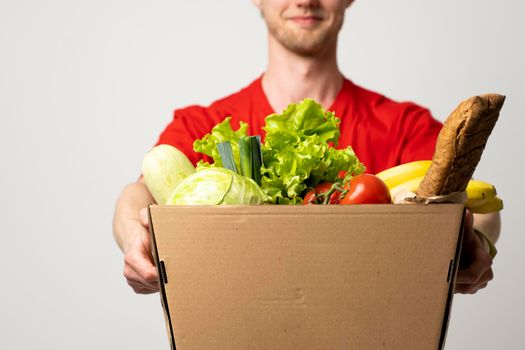 Food delivery service. Portrait of pleased delivery man in red uniform smiling while carrying paper box with food products isolated over white background