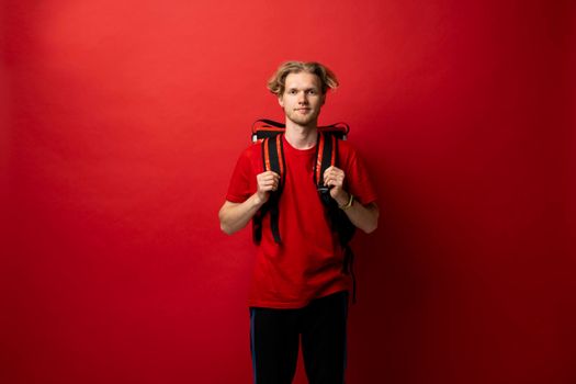 Young courier in a red uniform t-shirt and with red food thermo bag on a shoulder standing isolated on red background studio. Food delivery service