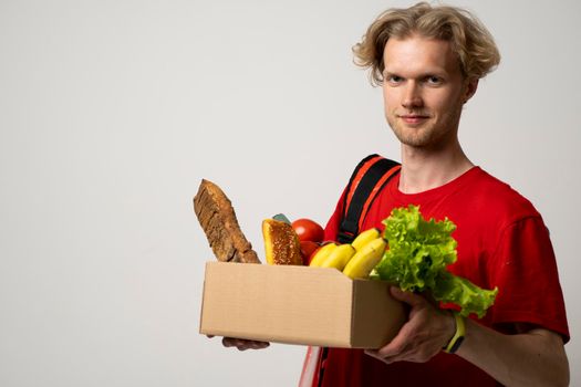 Portrait of smiling handsome delivery man holding paper grocery box with a groceries isolated on white background