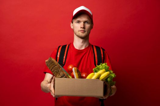 Delivery male employee in a red uniform holds paper cardbox package with groceries on a red background. Products delivery from shop or restaurant to home. Copy space