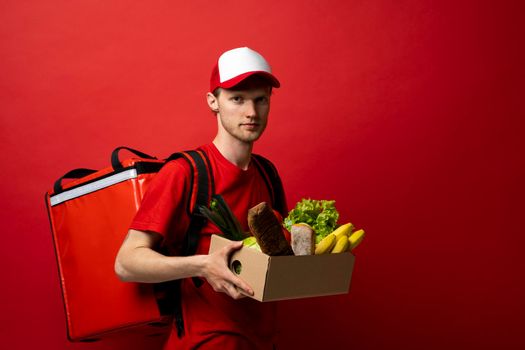 Delivery man in red uniform holds craft cardboard box with food isolated on white background, studio portrait. Service concept