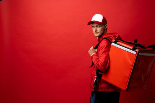 Portrait of a young courier, delivery man in red uniform with a thermal backpack isolated on a red background. Fast home delivery. Online order. Courier delivers groceries home