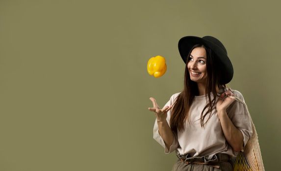 Smiling brunette young woman in a beige t-shirt and a hat holding a reusable mesh bag with fresh vegetables and fruits on a shoulder and throwing a yellow pepper in a air. Zero waste concept