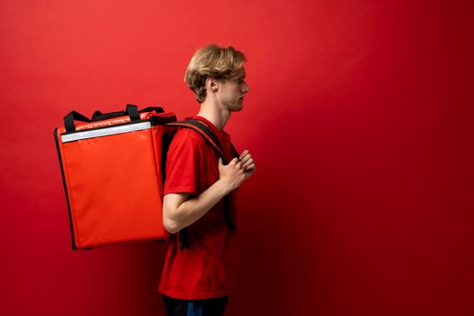 Side view of young courier in a red t-shirt and with a thermo bag on color red background, space for text. Food delivery service