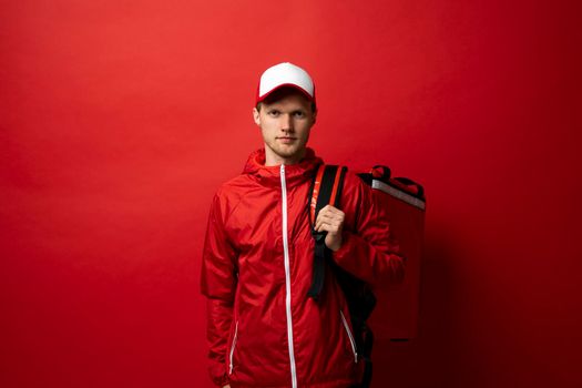 Young courier in a red uniform t-shirt and with red food thermo bag on a shoulder standing isolated on red background studio. Food delivery service