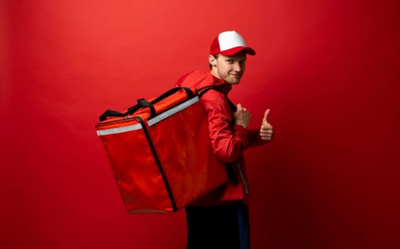 Delivery guy employee man in red cap and uniform workwear work as dealer courier hold red thermal food bag and showing thumb up isolated on red color background studio. Service concept