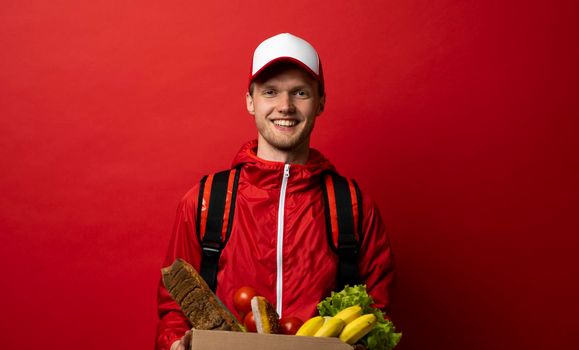 Portrait of smiling handsome delivery man holding paper grocery box with a groceries isolated on red background