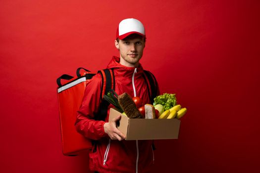 Delivery man in red uniform holds cardboard paper box with food isolated on white background, studio portrait. Service concept