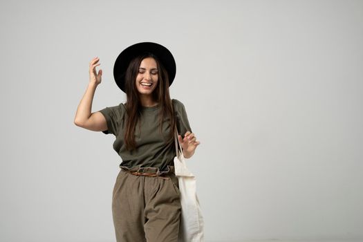 Brunette woman in green t-shirt and black hat holding cotton shopper bag with vegetables, products in white room. Eco friendly shopping bags. Zero waste, plastic free concept