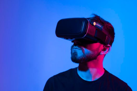 Portrait of man using VR headset while playing a video game in dark interior illuminated neon light. Close-up futuristic googles with colored light