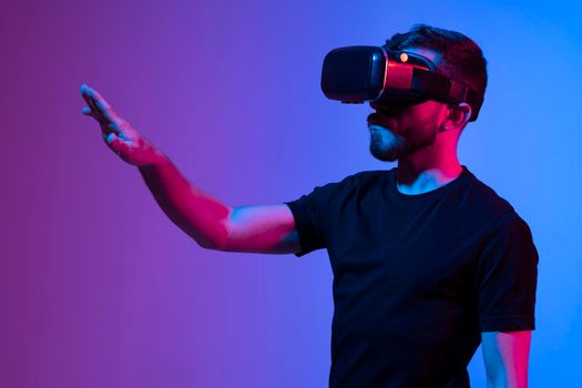 Portrait of man using VR headset while communicating with a friends in a metaverse in dark interior illuminated neon light. Close-up futuristic googles with colored light