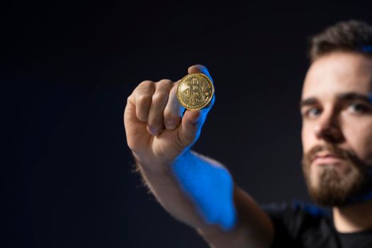 Mature bearded man wearing in black t-shirt holding gold bitcoin in hand and standing isolated on black background