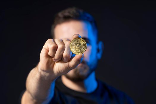 Mature bearded man wearing in black t-shirt holding gold bitcoin in hand and standing isolated on black background