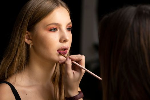 Makeup artist applies red lipstick on a beautiful woman face. Hand of make-up master, painting lips of young beauty model girl. Make up in process