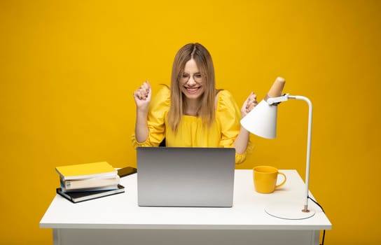 Excited young woman working with a grey laptop computer, notebook while sitting at the table with grey laptop. Smiling business woman or student received a good news isolated on a yellow background