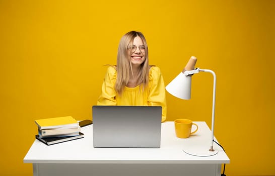 Excited young woman working with a grey laptop computer, notebook while sitting at the table with grey laptop. Smiling business woman or student received a good news isolated on a yellow background