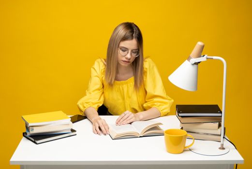 Tired young student woman in yellow casual clothes sitting at the table reading the book in library of university or college. Sitting and reading on yellow background. Studying