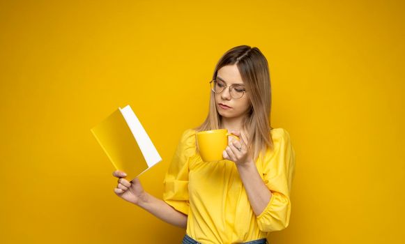 Beautiful smart young girl holding and reading book isolated on the yellow background. Portrait of attractive woman in a yellow blouse and wearing glasses reading book. Education, studying, knowledge