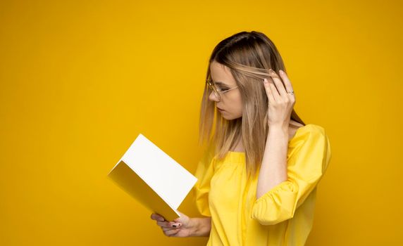 Portrait of a young student woman in a yellow t-shirt and glasses reading a book isolated over yellow background. Girl reading novel. Studying