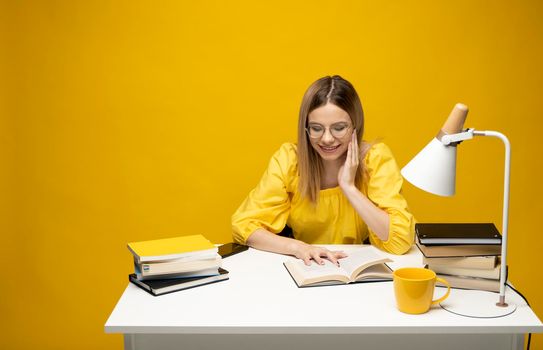 Young happy smiling student woman in yellow casual clothes reading the book at the table in library of university or college. Studying