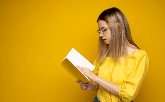 Beautiful smart young girl holding and reading book isolated on the yellow background. Portrait of attractive woman in a yellow blouse and wearing glasses reading book. Education, studying, knowledge