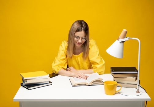 Young happy smiling student woman in yellow casual clothes reading the book at the table in library of university or college. Studying