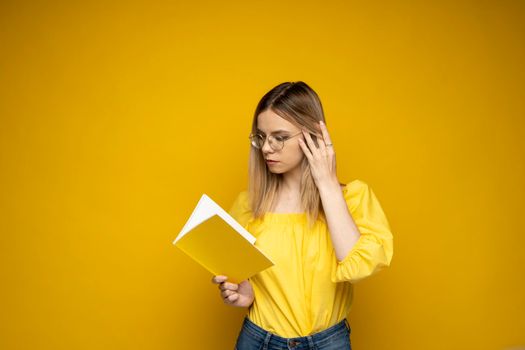 Beautiful smart young girl holding and reading book isolated on the yellow background. Portrait of attractive woman in a yellow blouse and wearing glasses reading book. Education, studying, knowledge