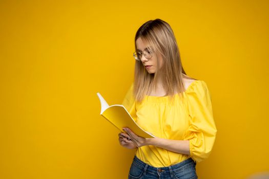 Beautiful smart young girl holding and reading book isolated on the yellow background. Portrait of attractive woman in a yellow blouse and wearing glasses reading book. Education, studying, knowledge