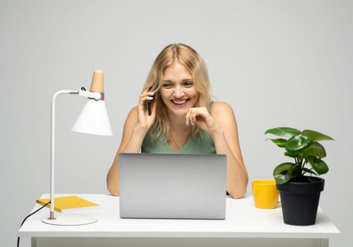 Young attractive business woman with a blond hair sitting at the table and working on a laptop and talking with a client on the phone. Young cheerful student girl talking with a smartphone. Studying