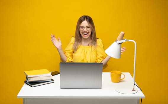 Excited young woman working with a grey laptop computer, notebook while sitting at the table with grey laptop. Smiling business woman or student received a good news isolated on a yellow background