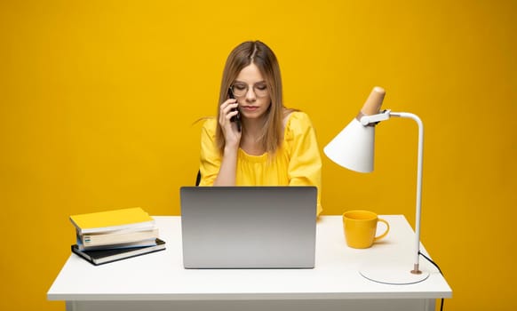 Young attractive business woman with a blond hair sitting at the table and working on a laptop and talking with a client on the phone. Young cheerful student girl talking with a smartphone. Studying