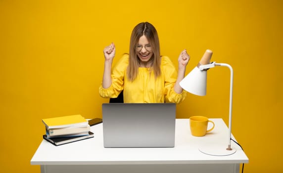 Excited young woman working with a grey laptop computer, notebook while sitting at the table with grey laptop. Smiling business woman or student received a good news isolated on a yellow background