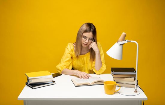 Tired young student woman in yellow casual clothes sitting at the table reading the book in library of university or college. Sitting and reading on yellow background. Studying