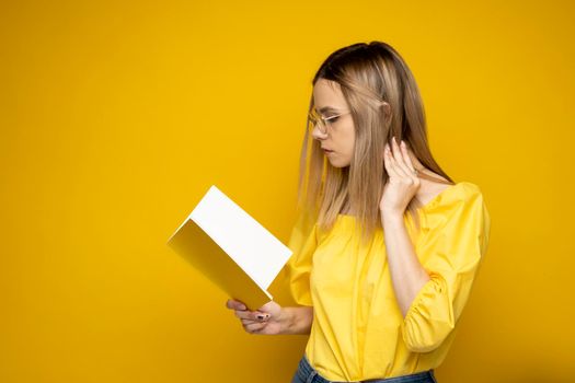 Beautiful smart young girl holding and reading book isolated on the yellow background. Portrait of attractive woman in a yellow blouse and wearing glasses reading book. Education, studying, knowledge