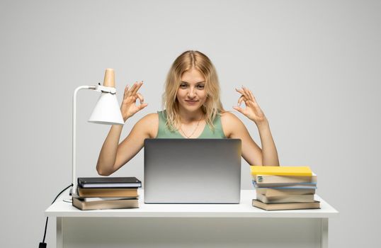 Student girl showing ok gesture with both hands, happy with completed work, smiling and looking at the camera while working on laptop computer sitting in front of big piles of books