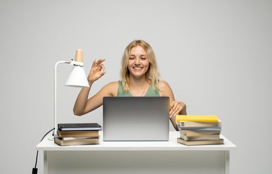 Student girl showing ok gesture with both hands, happy with completed work, smiling and looking at the camera while working on laptop computer sitting in front of big piles of books