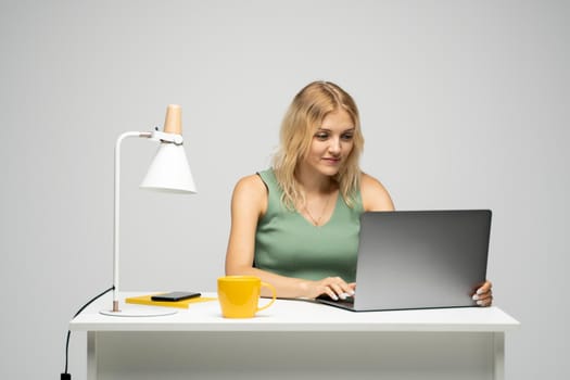 Smiling business woman working with a laptop isolated on a grey background. Portrait of a pretty young woman studying while sitting at the table with grey laptop computer, notebook.