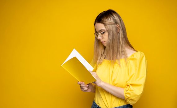 Young teenage woman in a yellow t-shirt and glasses reading a book isolated over yellow background. Girl reading novel. Studying