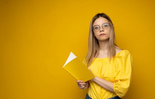 Beautiful smart young girl holding and reading book isolated on the yellow background. Portrait of attractive woman in a yellow blouse and wearing glasses reading book. Education, studying, knowledge