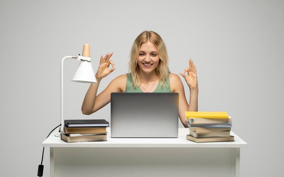 Student girl showing ok gesture with both hands, happy with completed work, smiling and looking at the camera while working on laptop computer sitting in front of big piles of books