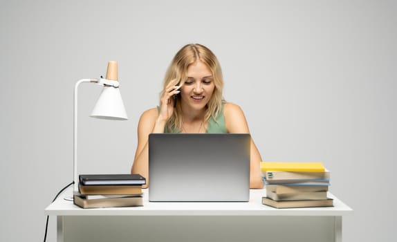 Young attractive business woman with a blond hair sitting at the table and working on a laptop and talking with a client on the phone. Young cheerful student girl talking with a smartphone. Studying