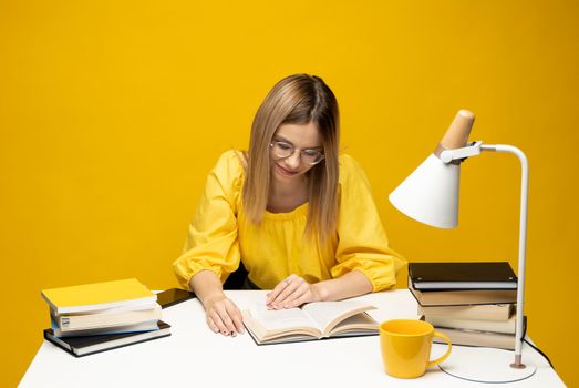 Young happy smiling student woman in yellow casual clothes reading the book at the table in library of university or college. Studying