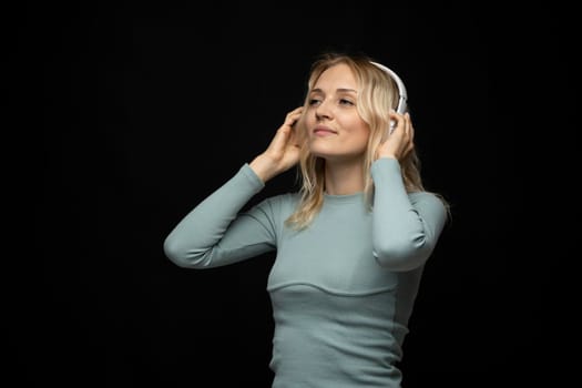 Beautiful attractive young blond woman wearing blue t-shirt and glasses in white headphones listening music and smiling on black background in studio. Relaxing and enjoying. Lifestyle