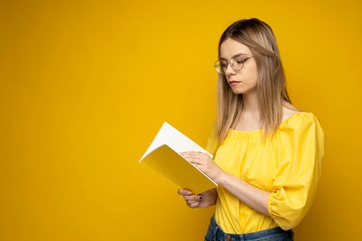 Beautiful smart young girl holding and reading book isolated on the yellow background. Portrait of attractive woman in a yellow blouse and wearing glasses reading book. Education, studying, knowledge